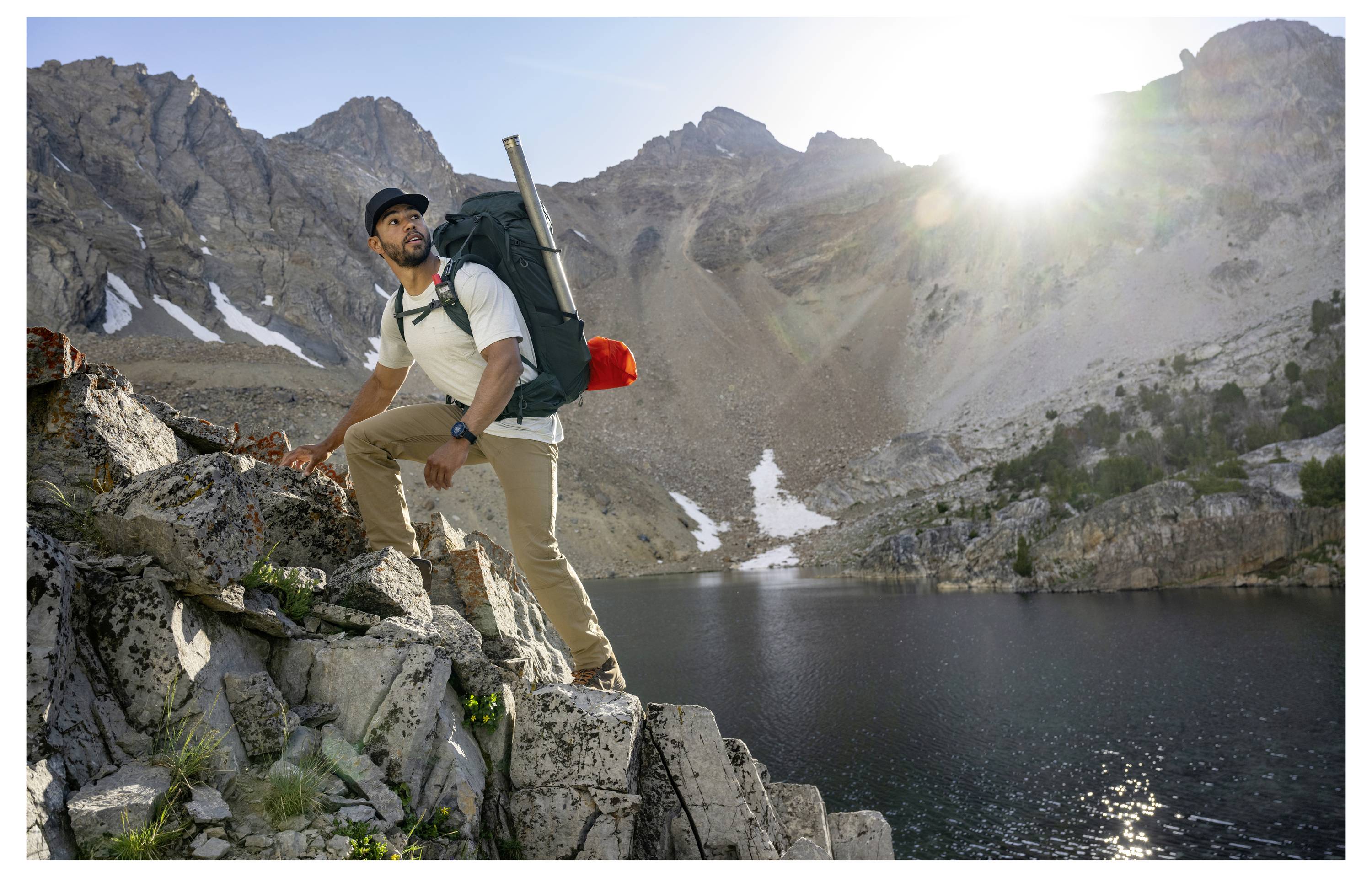 A person wearing hiking gear stands on rocky terrain by a lake, with a mountainous backdrop and sunlight streaming over peaks.