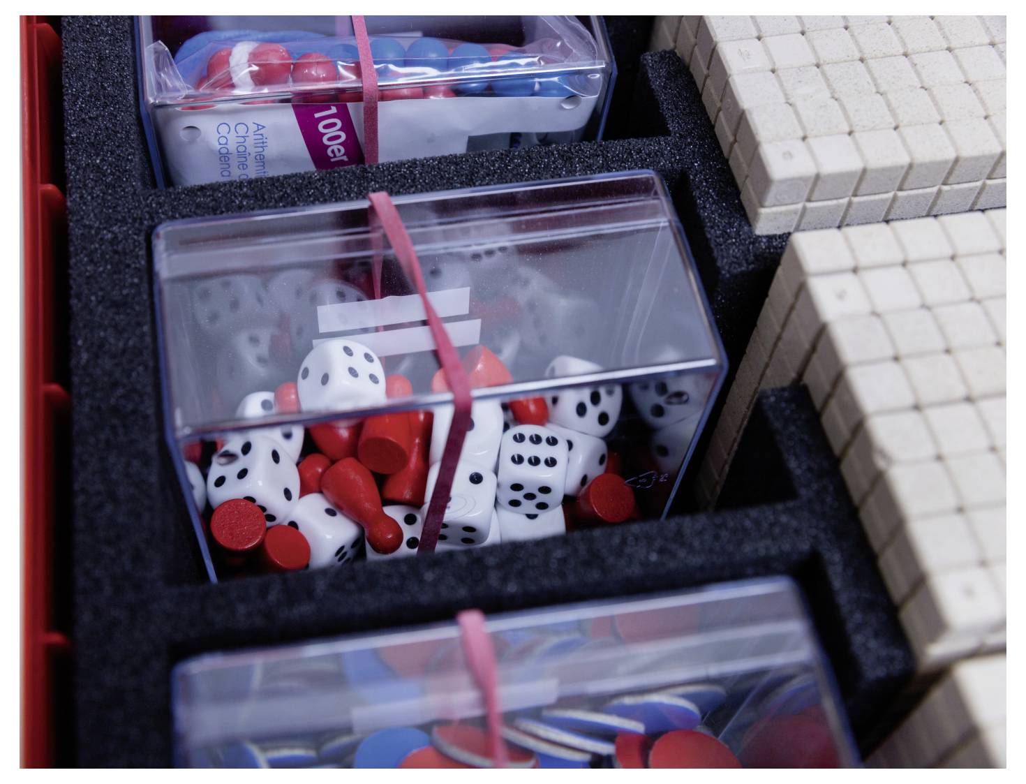 A box filled with educational math tools, including dice, red and white chips, and stacked wooden blocks, organized in compartments.