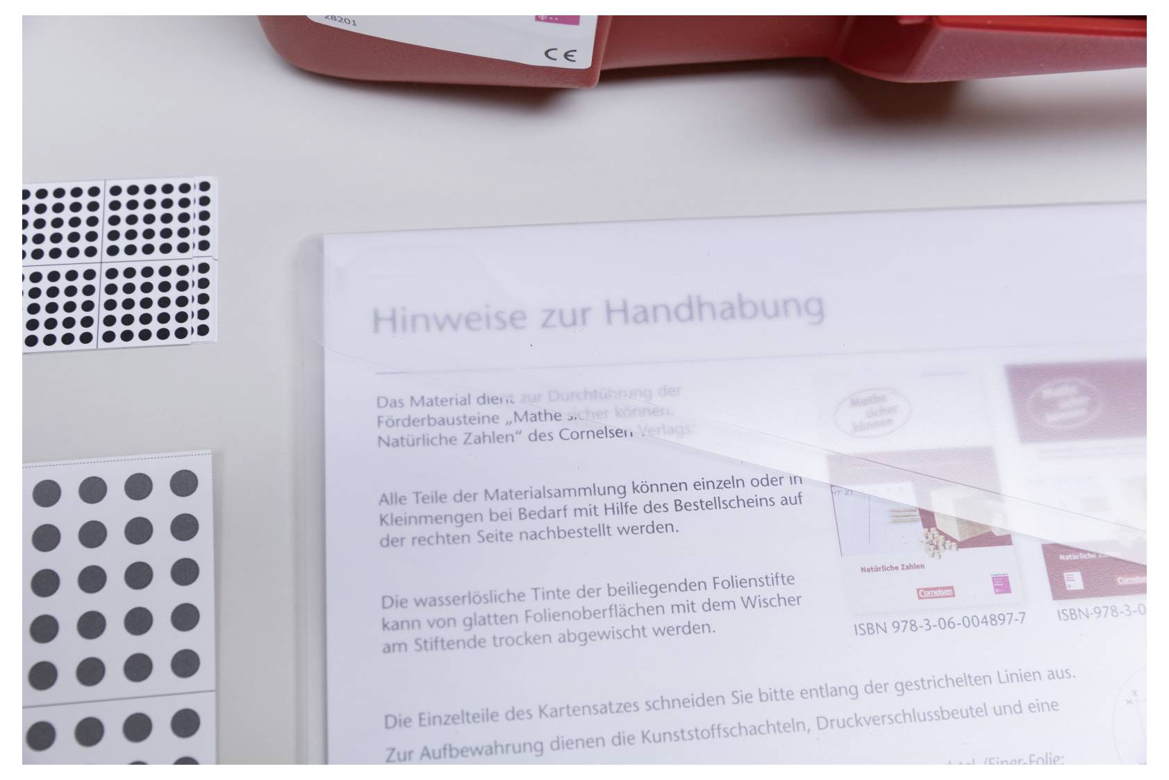 'Hinweise zur Handhabung' document in German on a desk with stickers and a box. Text discusses material usage and storage instructions.