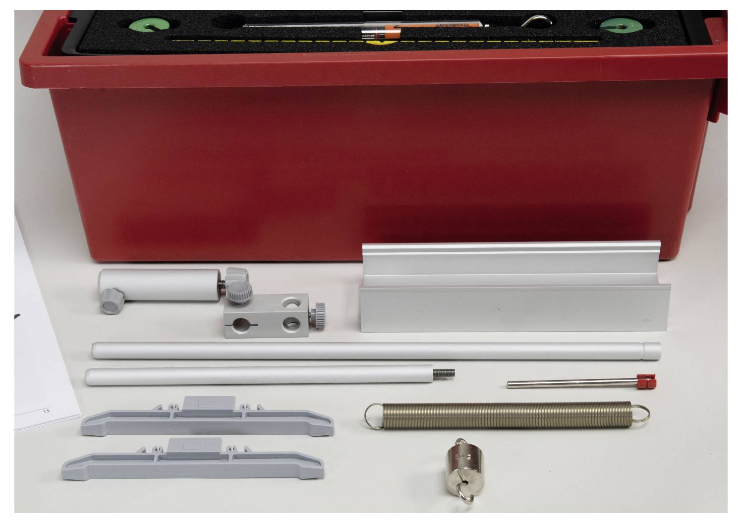 A red storage box contains assembly parts including rods, brackets, a ruler, and a cylindrical weight, organized on a white surface.