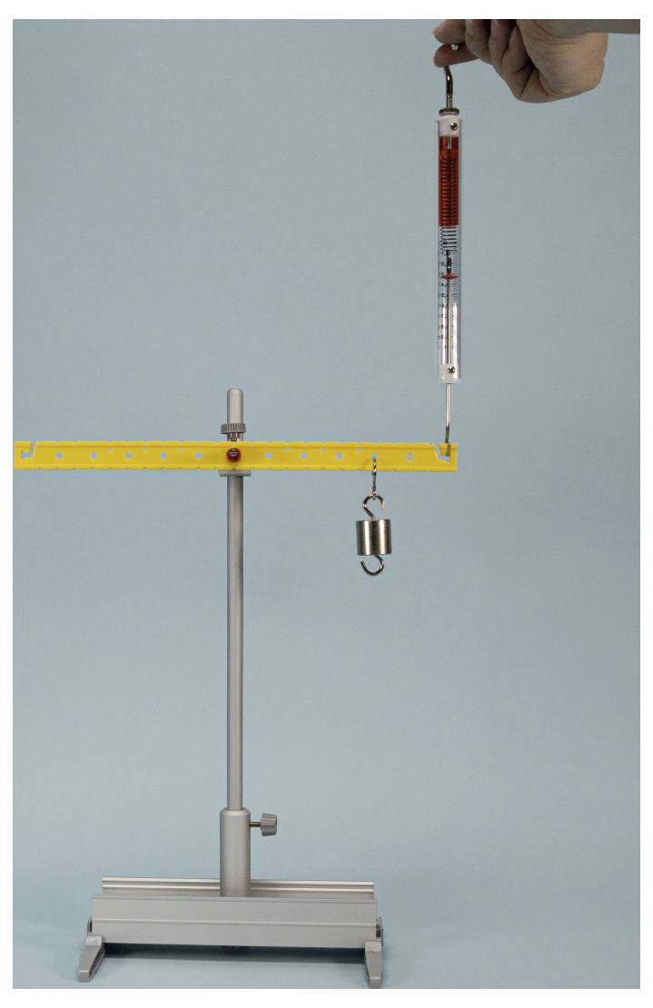 A hand holds a spring scale attached to a hanging weight on a stand against a light blue background, illustrating force measurement.
