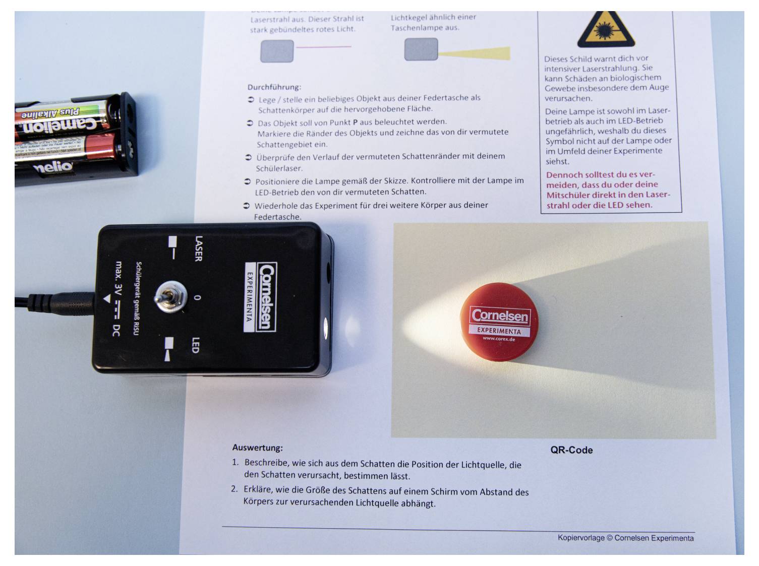 A device projects light onto a round red object labeled 'Cornelsen Experimenta'. Instruction sheet and batteries are visible nearby.
