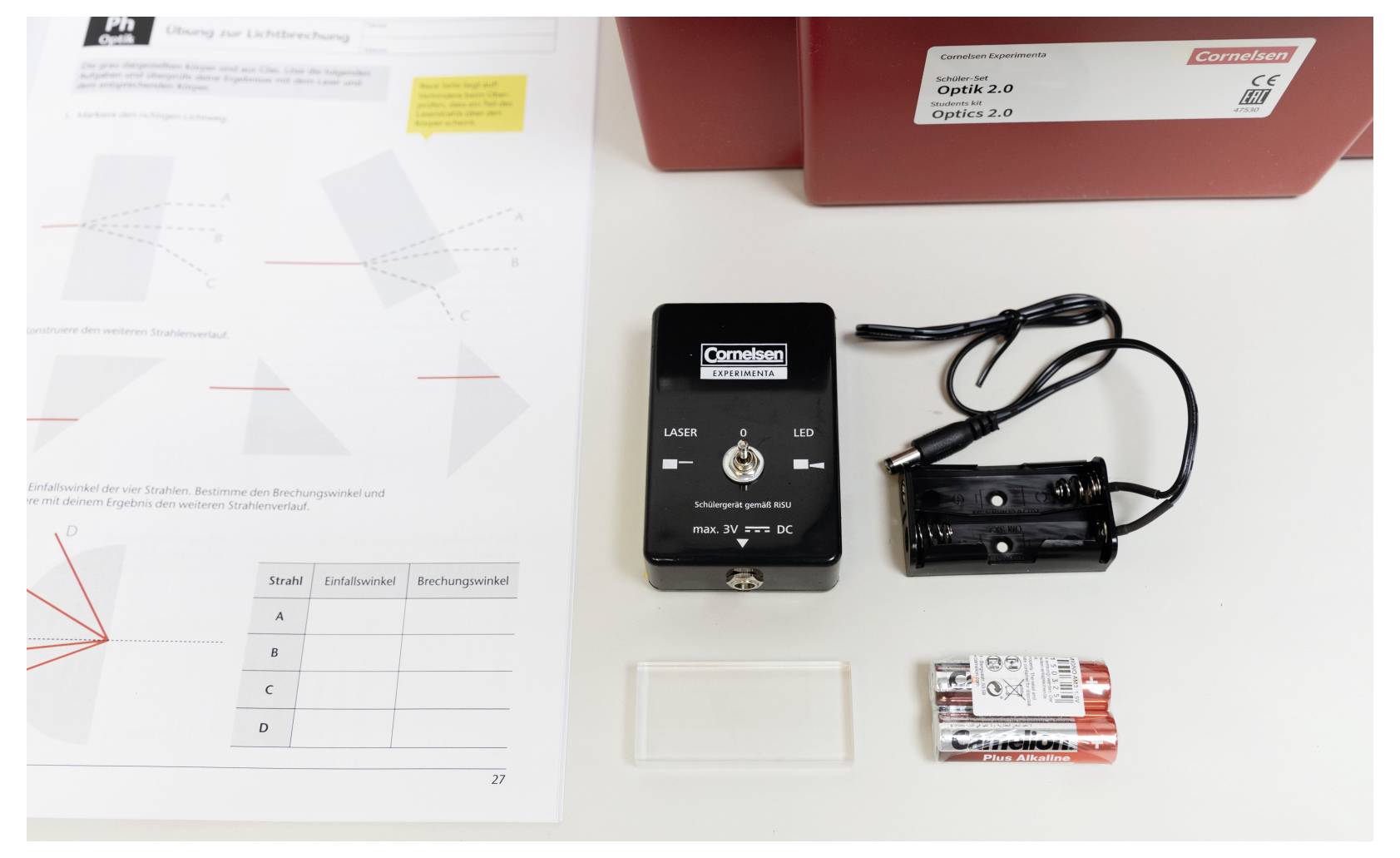 'A desk with a laser module, battery holder with AA batteries, a small rectangular object, and an optics worksheet labeled "Optik 2.0".'