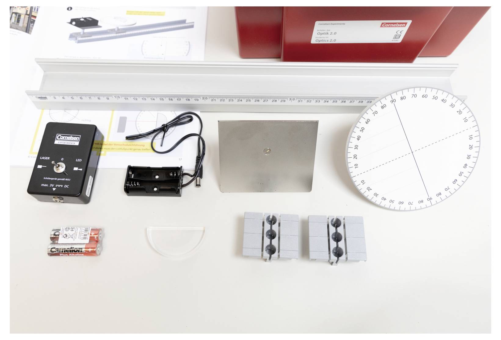 A metal ruler, angle measuring device, battery holder, metal plate, plastic components, batteries, and red boxes on a table.