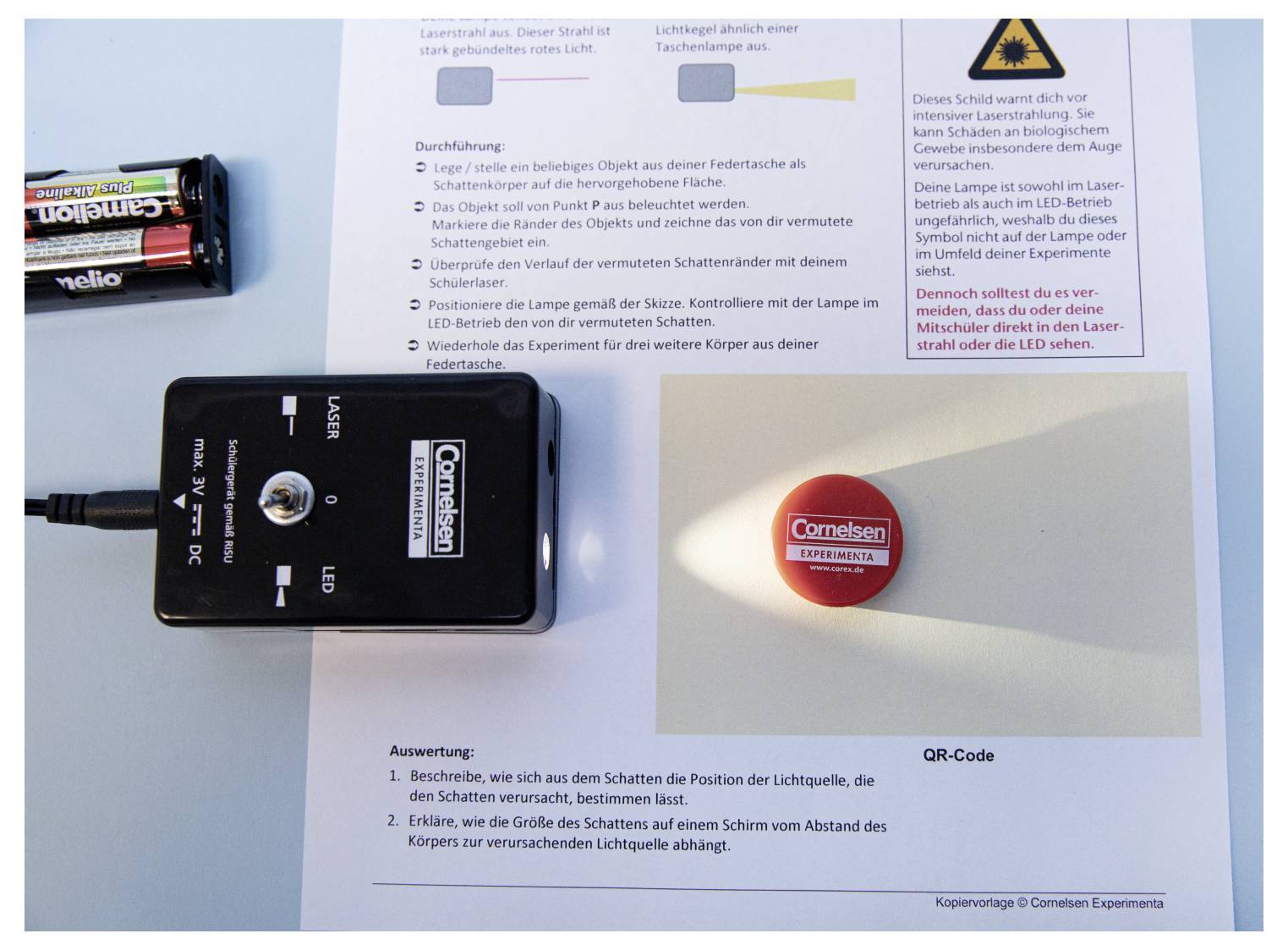 A small LED light shines on a red button marked 'Cornelsen', casting a shadow on a paper with text and diagrams, beside two batteries.