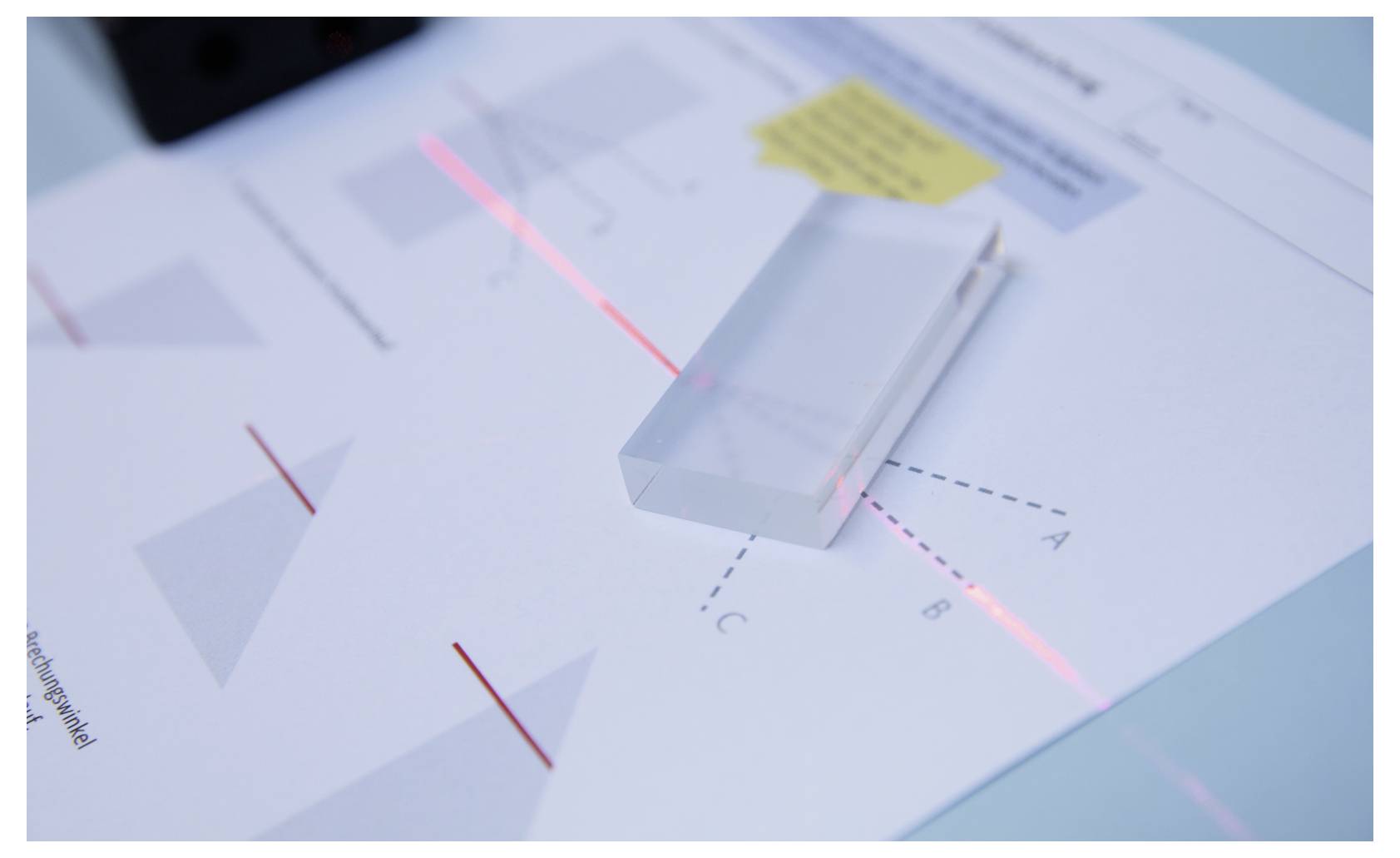 A rectangular prism on a paper with geometric diagrams shows a refraction experiment, as a red laser passes through it, bending light.