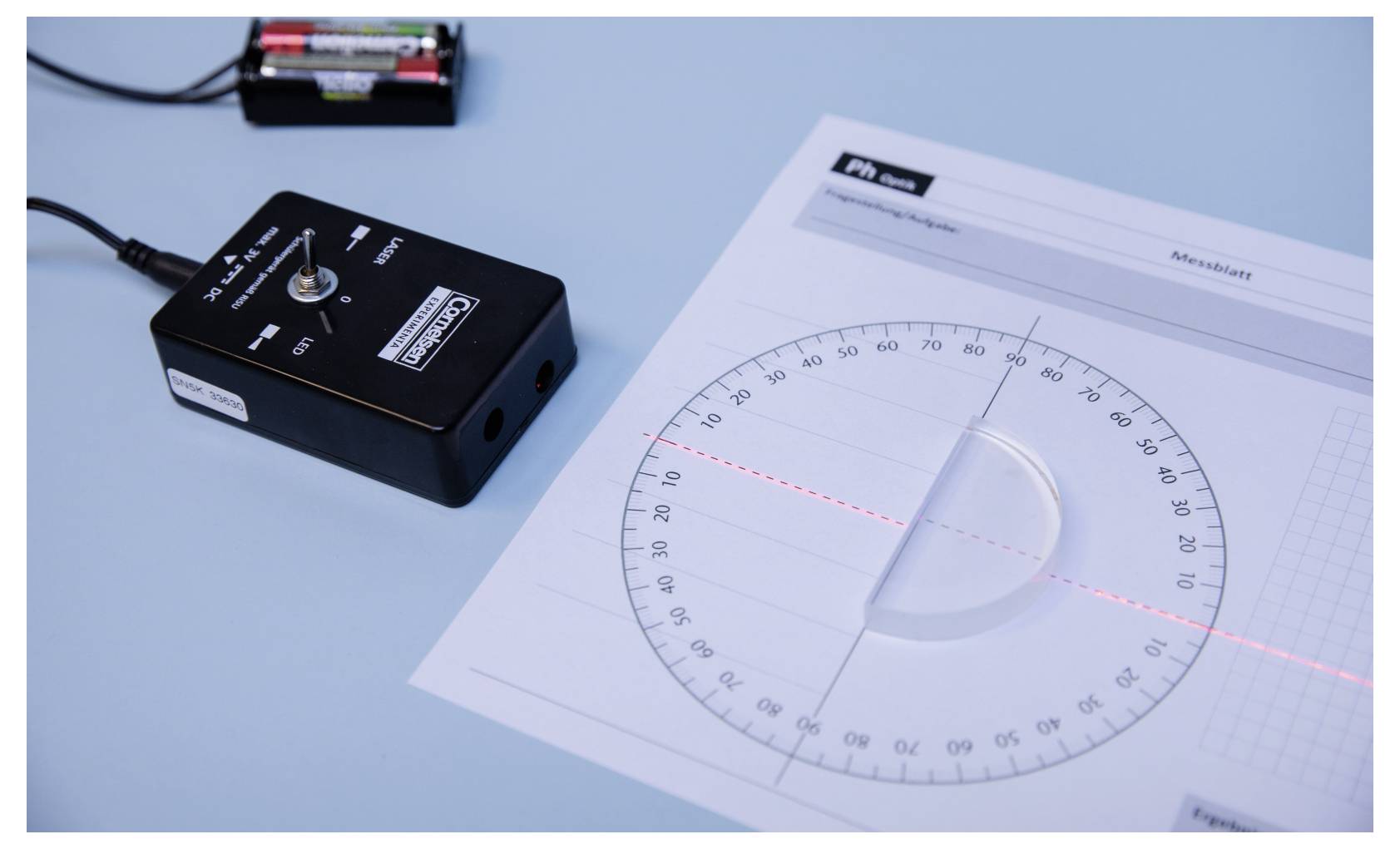 'A protractor with a laser beam intersects the center, highlighting angles. Nearby, a power unit with connected wires is visible on a blue surface.'