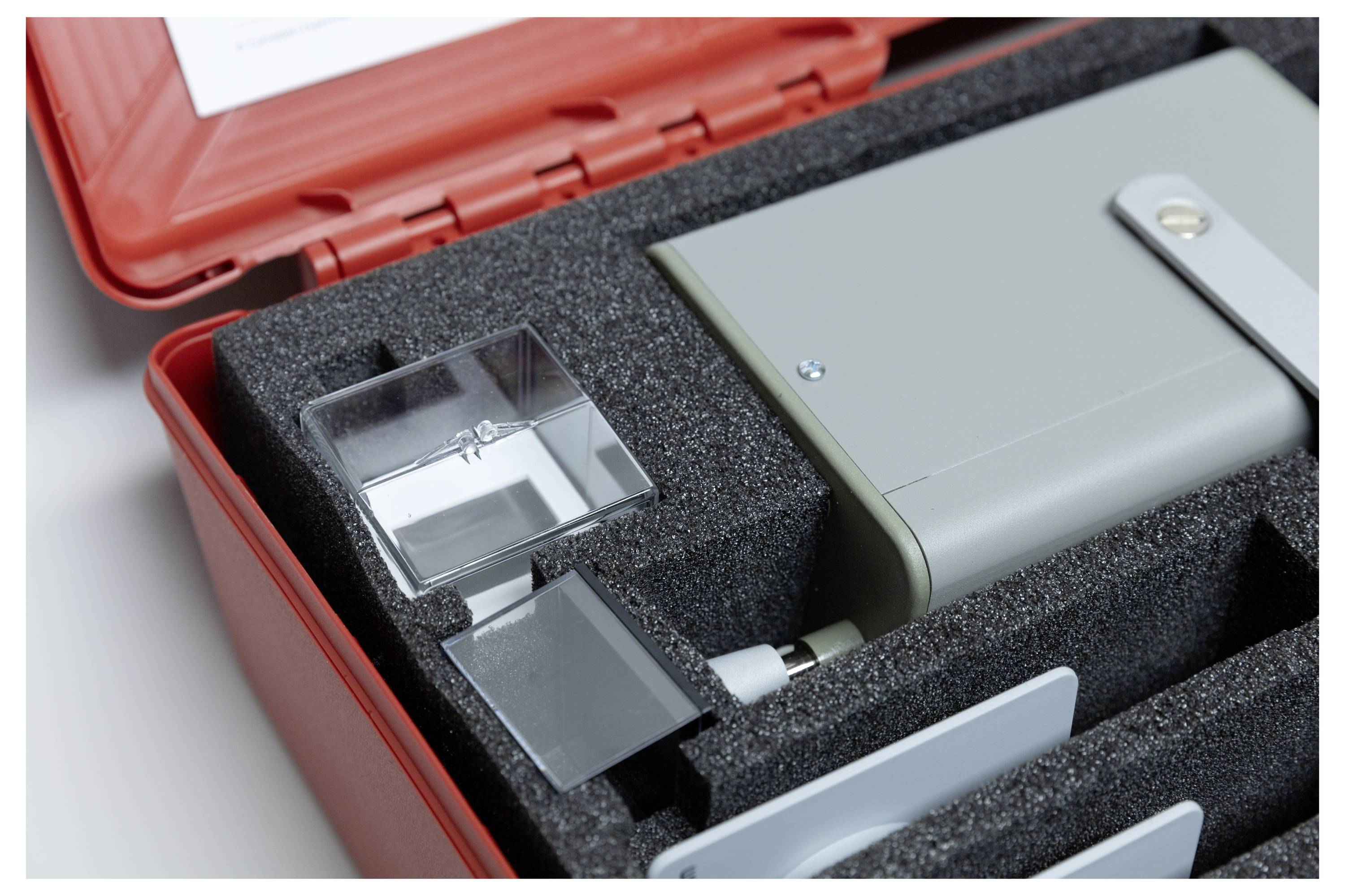 A gray electronic device with a metal latch resting in a foam-lined red case, next to a clear glass block, potentially equipment for analysis or measurement.