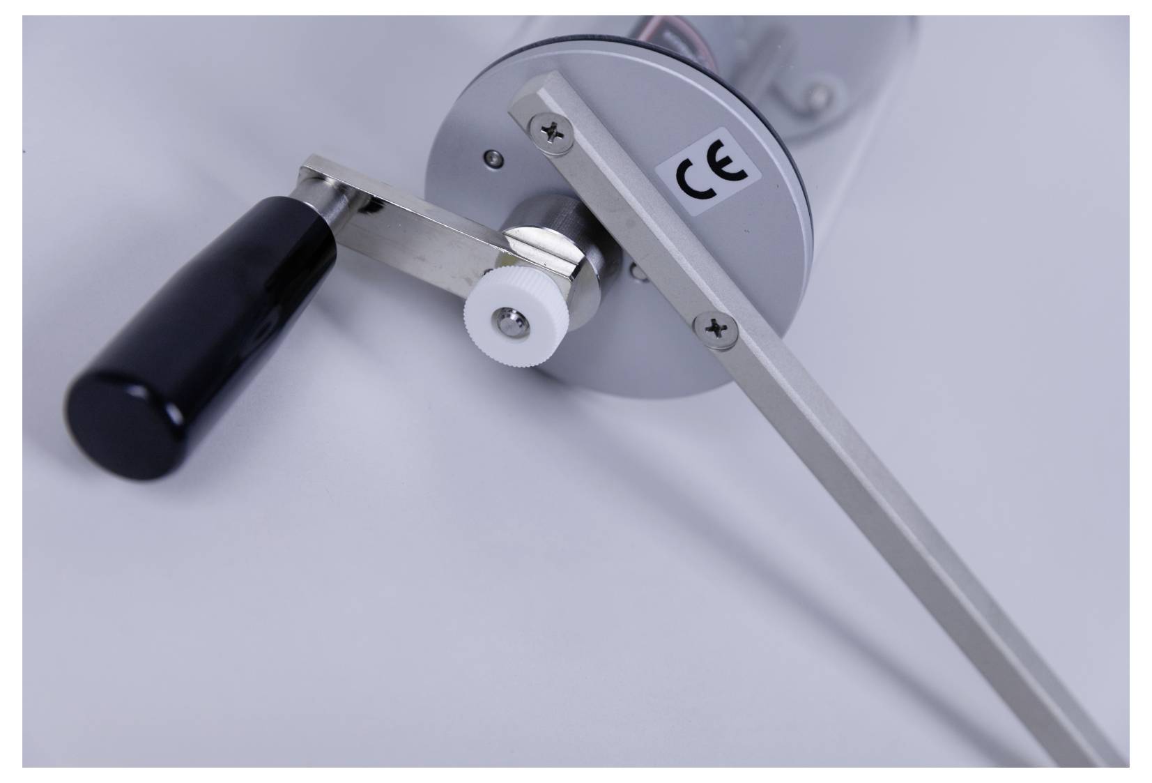 Manual crank handle connected to a machine part, featuring a black grip and visible 'CE' marking, indicating compliance with safety standards.