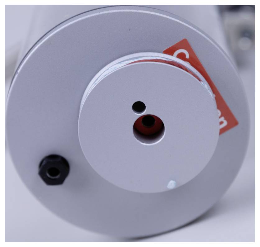 A close-up of a cylindrical metal device with a red sticker, displaying two small holes on the front. A black connector is visible on the side.