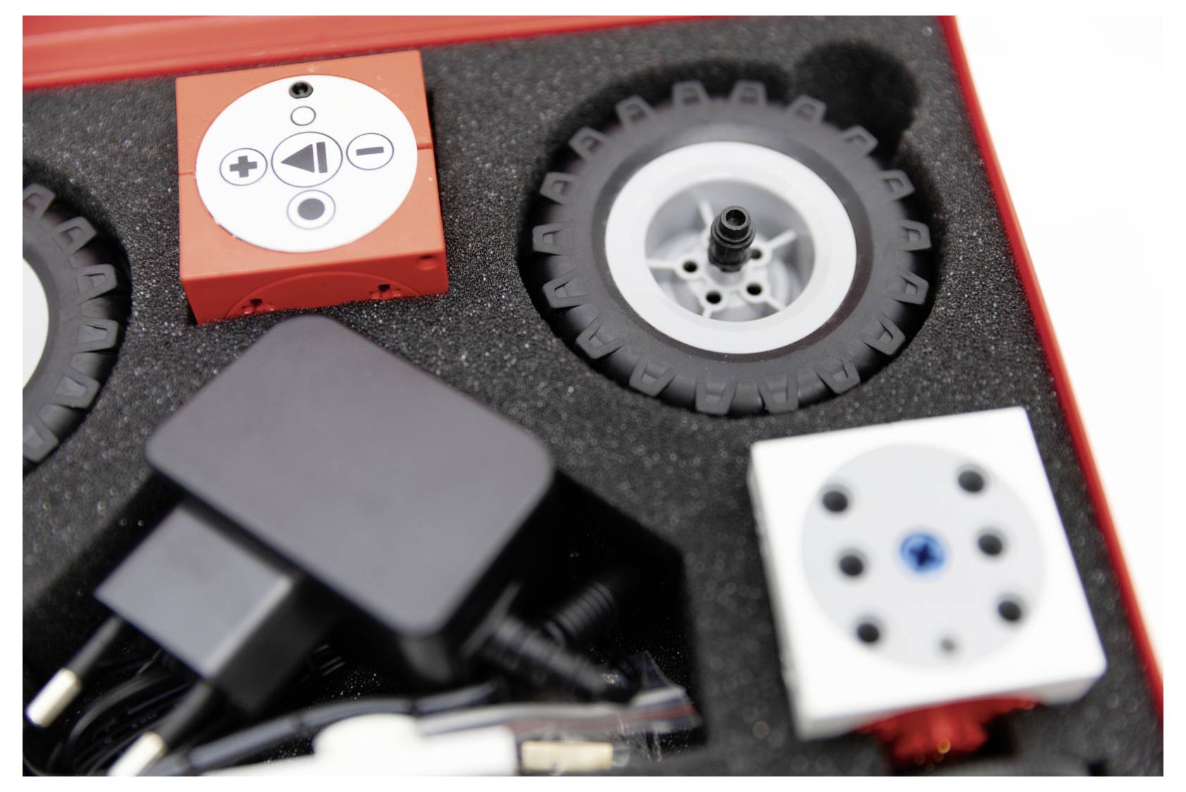 A foam-lined box containing a toy wheel, a red block with play controls, a white block, and an electronic adapter.