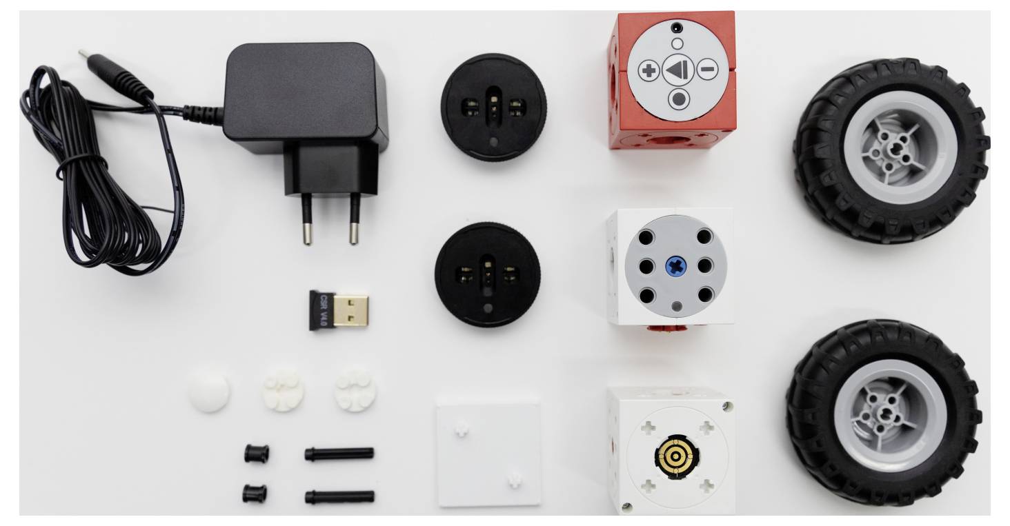 Robot kit components on a white surface, including wheels, connectors, charging plug, blocks with sensors, and USB adapter.