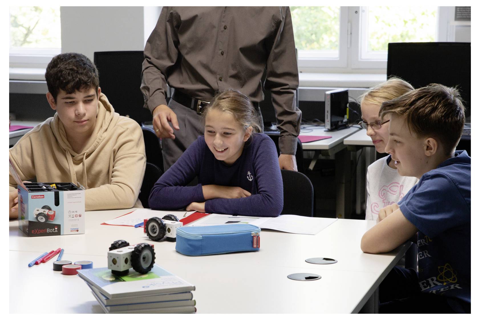 Students gathered around a table with robotics kits, smiling and discussing, guided by an adult in an educational setting.
