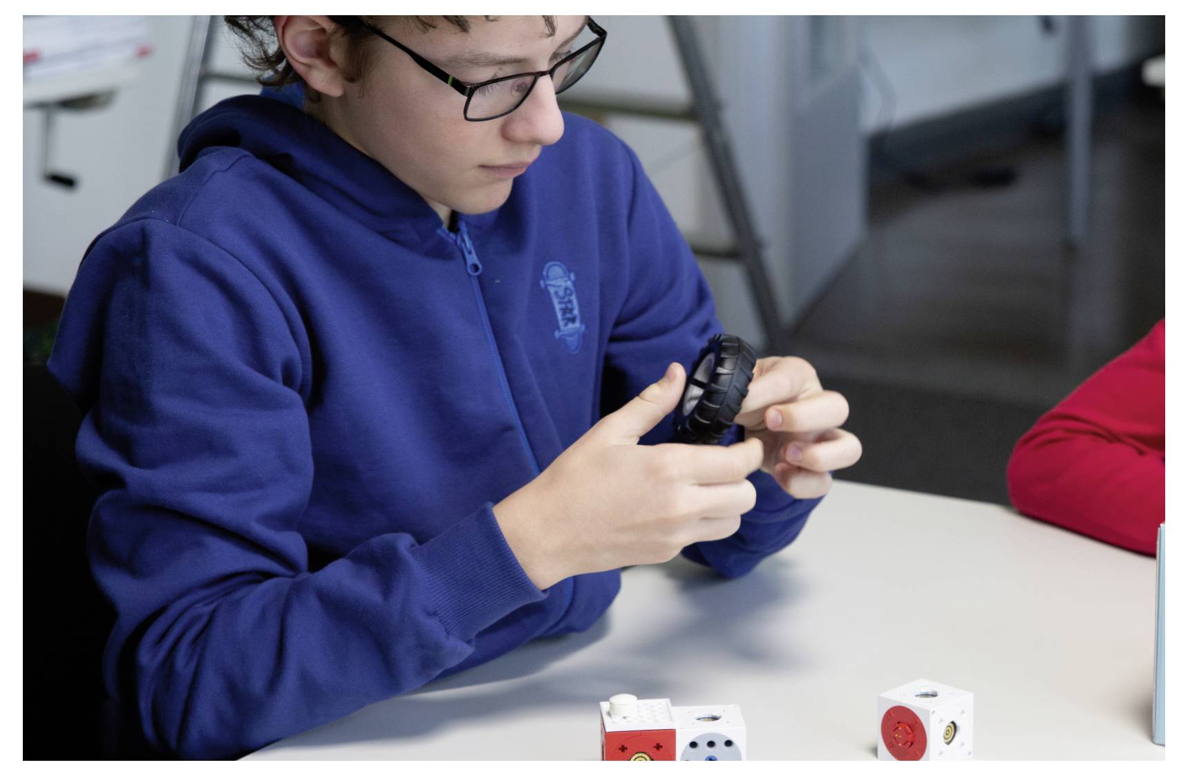 Boy in a blue hoodie examines a black gear-like object while sitting at a table with various small devices, focusing intently.