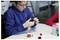 A person in a blue hoodie examines a robotic component at a table with white and red blocks, engaging in a STEM activity.