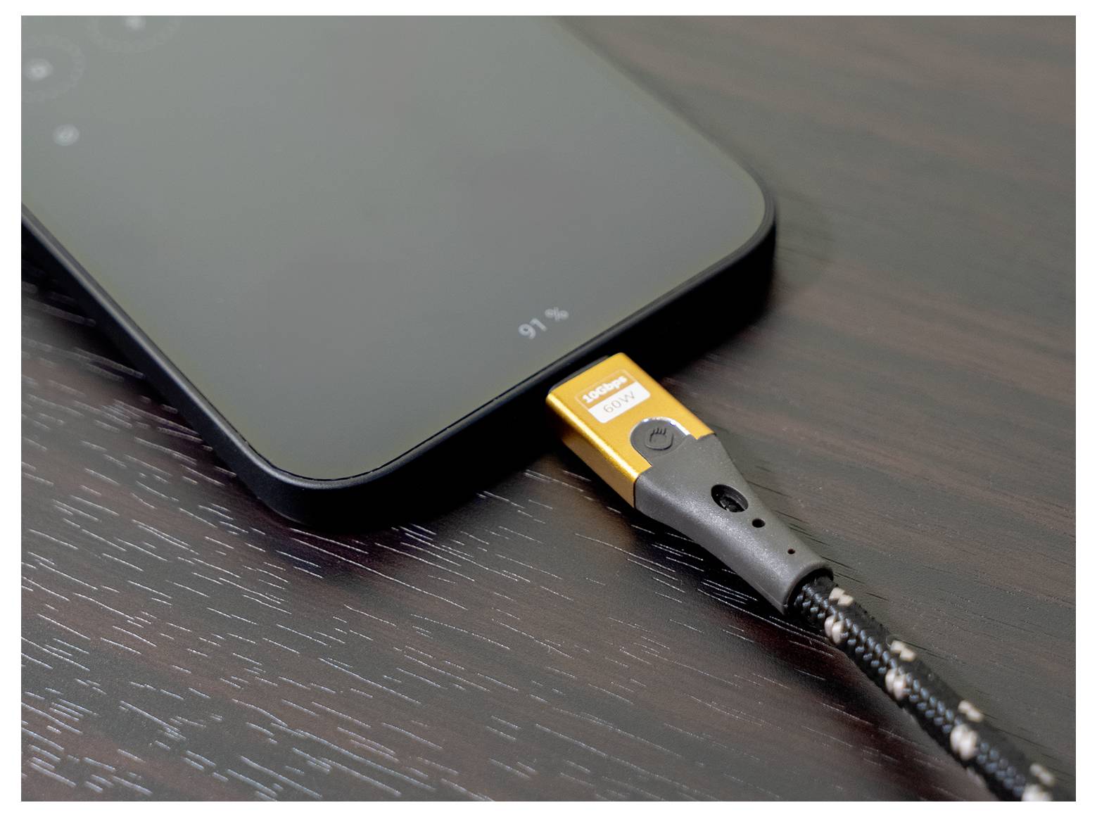 A smartphone charging on a wooden surface with a cable connected to the bottom port. The phone display shows a charging percentage.