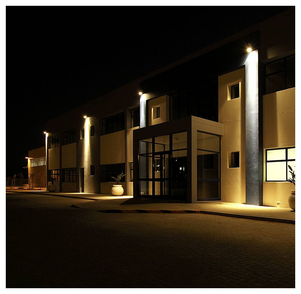 A modern building at night with illuminated exterior lights highlighting its structure, large glass entrance, and potted plants beside the doorway.