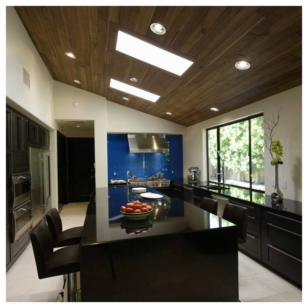 A modern kitchen with a high ceiling, wood paneling, and a large island featuring black countertops. Bright blue backsplash and natural light from a window.