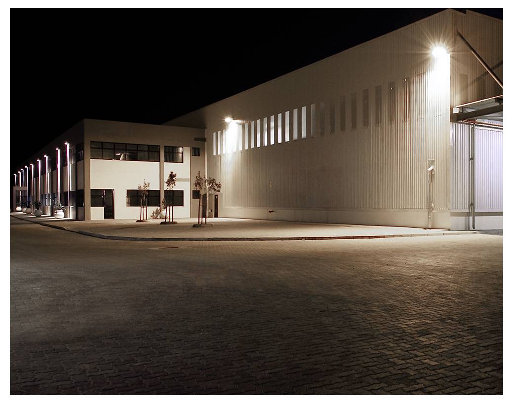 A large industrial building lit up at night, with bright exterior lighting casting shadows on the paved ground in an empty surroundings.