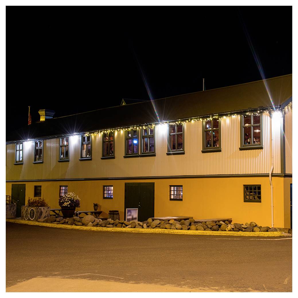 A two-story building with lit windows and strings of lights under a night sky. The exterior is painted yellow and white, and a sidewalk runs in front.