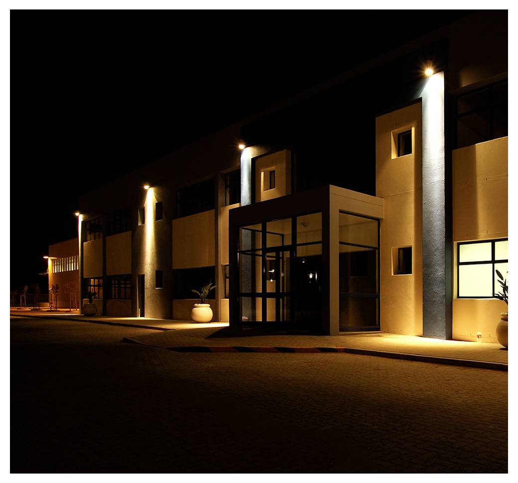 A modern building exterior at night, illuminated by soft lighting, with planters on either side of the entrance and large windows visible.