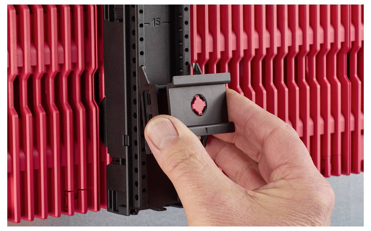 A hand attaches a black bracket to a red, ribbed panel on a wall, showing installation or assembly.