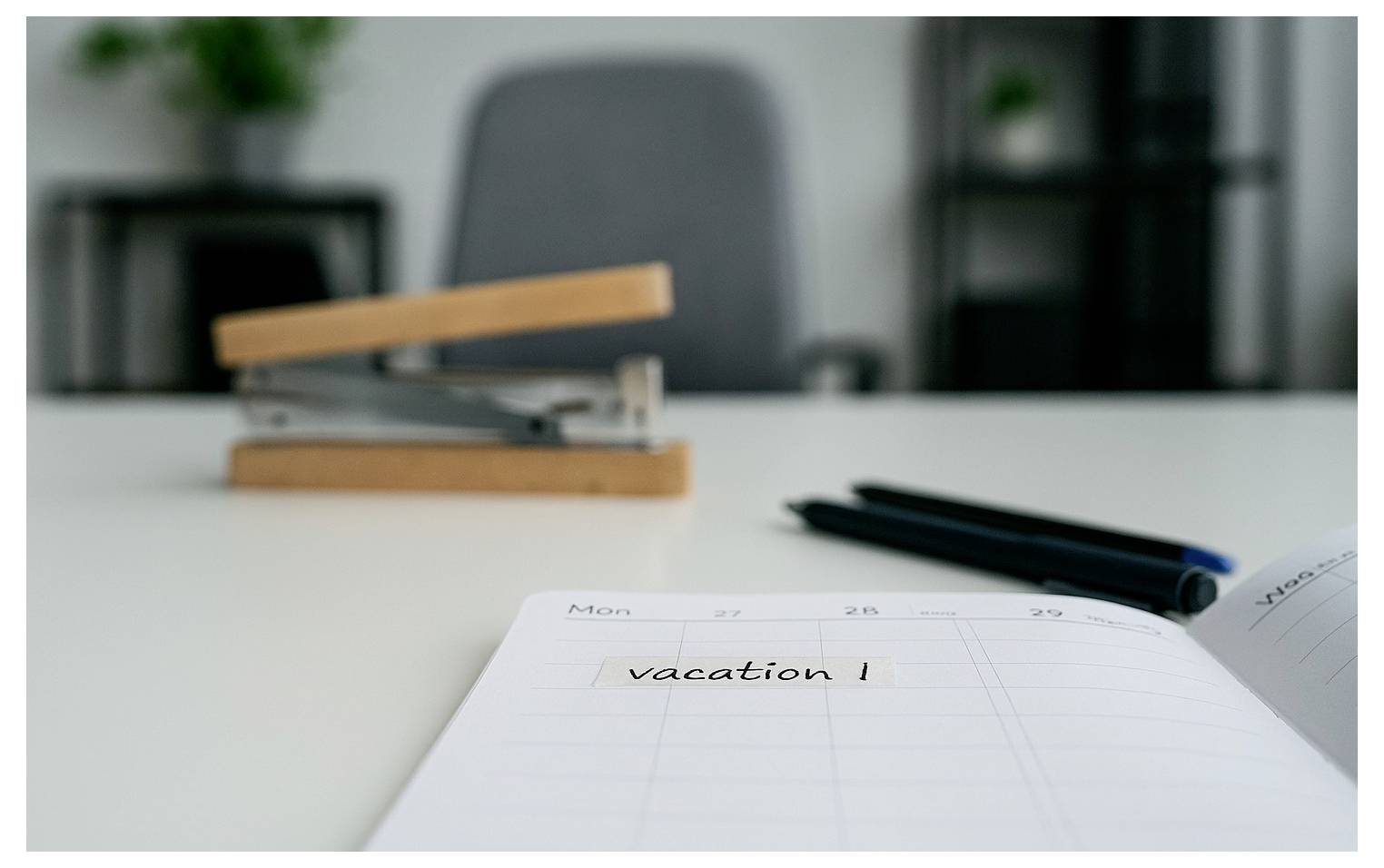 A desk with a stapler, pens, and an open planner displaying 'vacation!' on Monday. A blurred office chair and shelves are in the background.