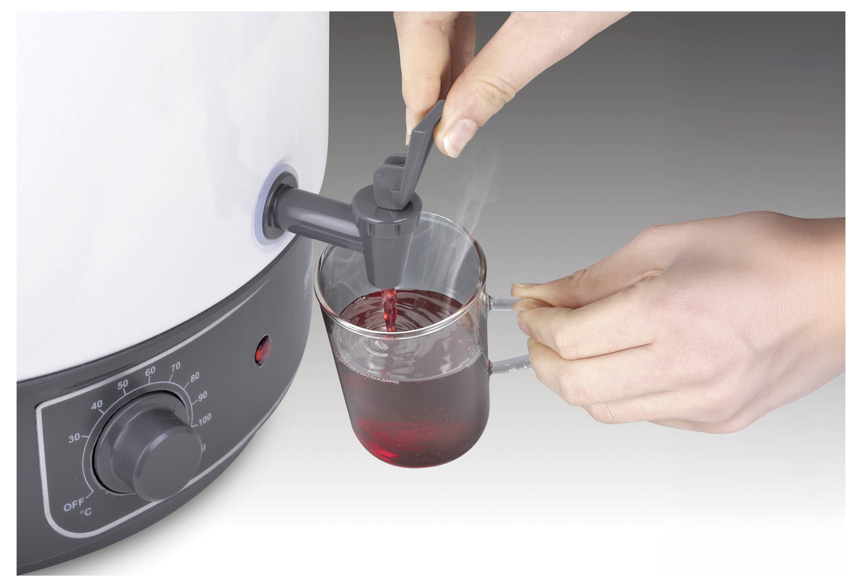 A person's hand dispenses hot beverage from a heated container into a clear mug. The container's dial is set to a middle temperature.