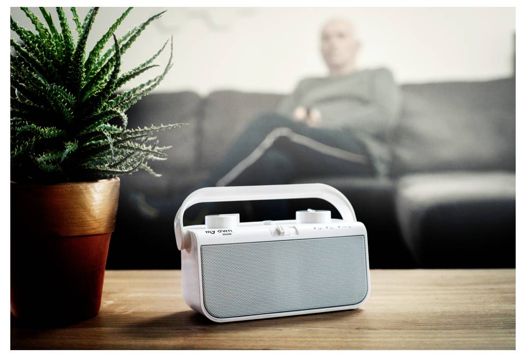 'A small white radio on a wooden table, a potted plant beside it; a person is sitting blurred in the background on a sofa.'