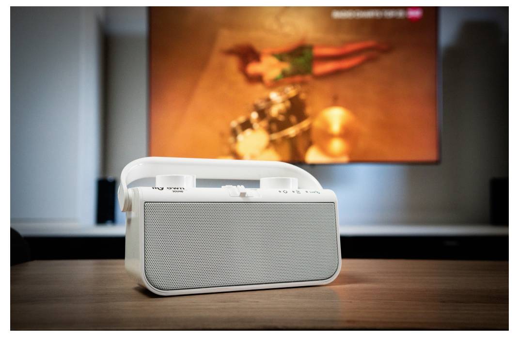 A white portable speaker on a wooden table with a blurry image of a person on a screen in the background.