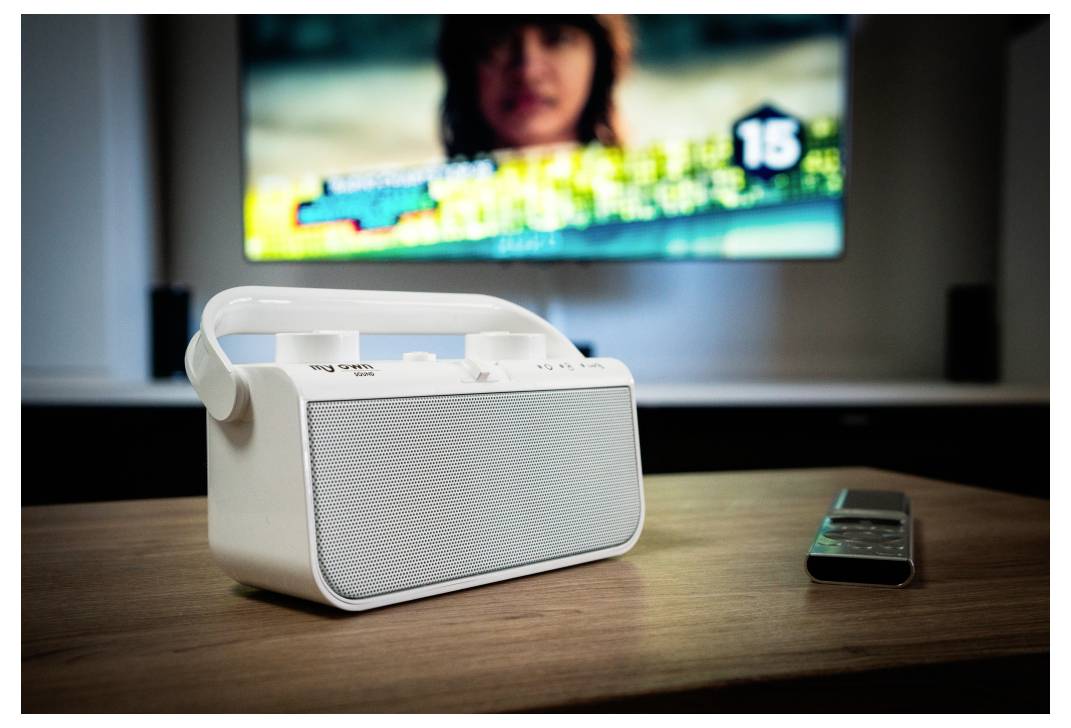 A white portable speaker with a handle sits on a wooden table, next to a remote control. A blurred TV screen is visible in the background.
