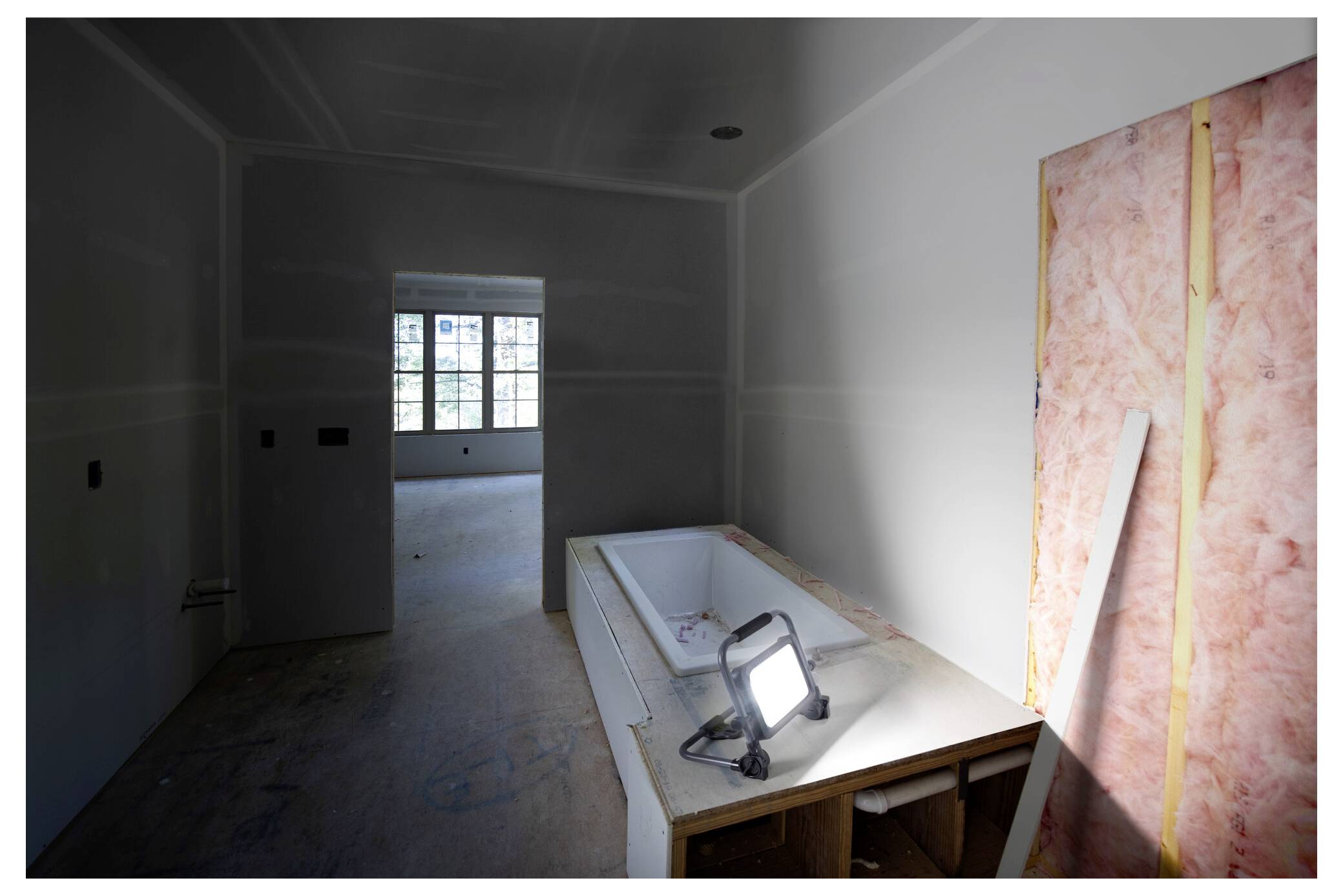'A room under construction with bare walls and floors, featuring a partially installed bathtub and a work lamp casting light.'