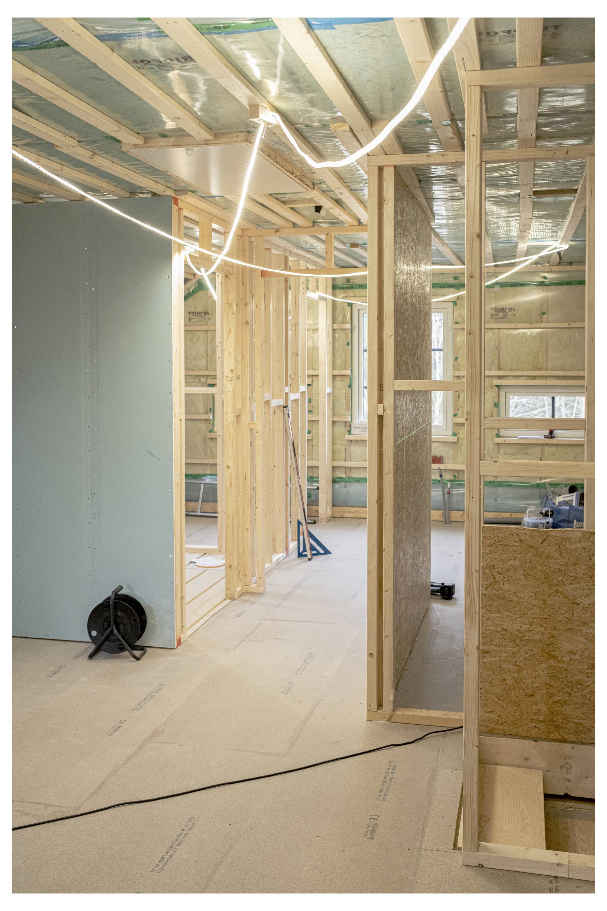 Construction site with wooden framing and insulation in progress, featuring partially installed drywall and exposed wiring overhead.