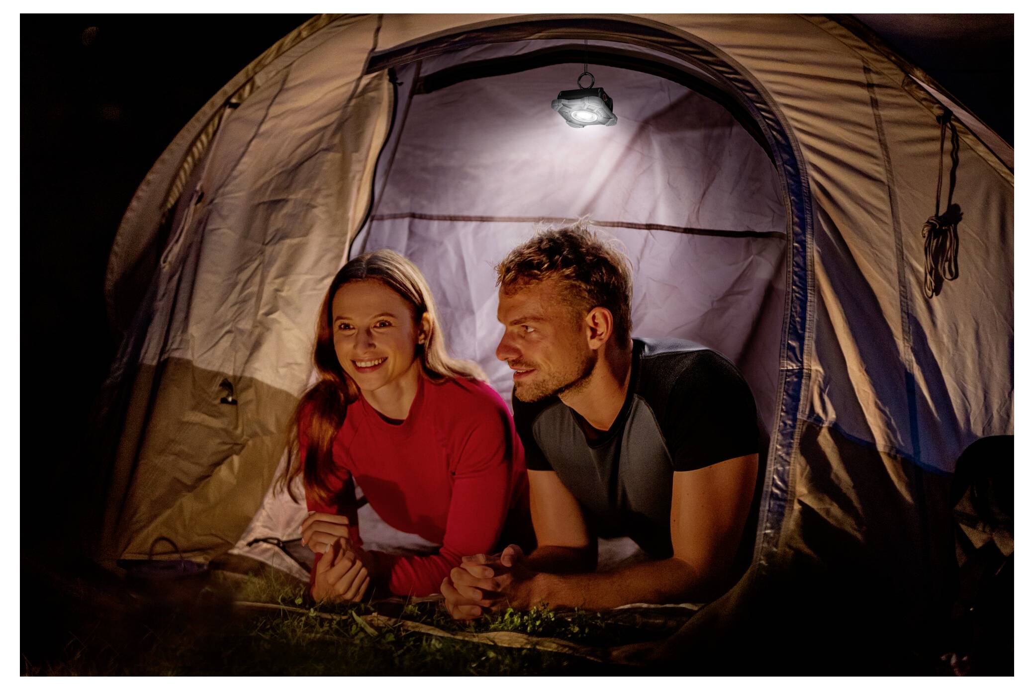 A couple lies inside a lit tent at night, smiling and talking, with camping gear visible outside. The scene suggests a relaxing outdoor adventure.