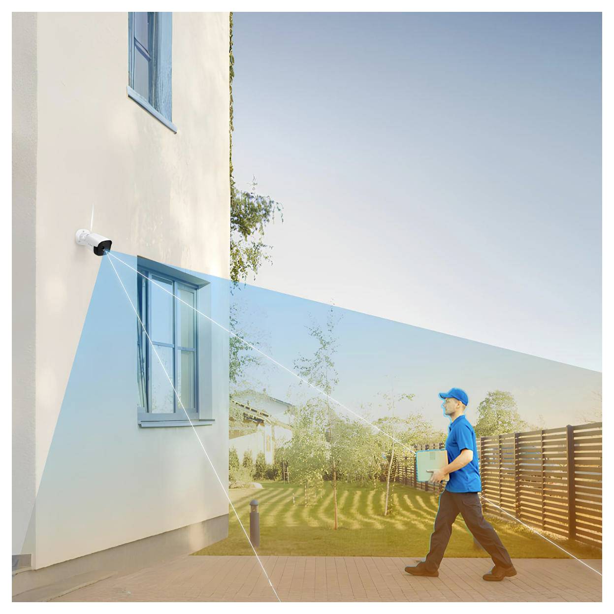 A security camera on a building wall monitors a man in a blue uniform carrying a package in a yard with trees and a fence.