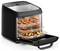 A black countertop air fryer oven with three trays filled with cooked chicken wings, sliced potatoes, and roasted vegetables.