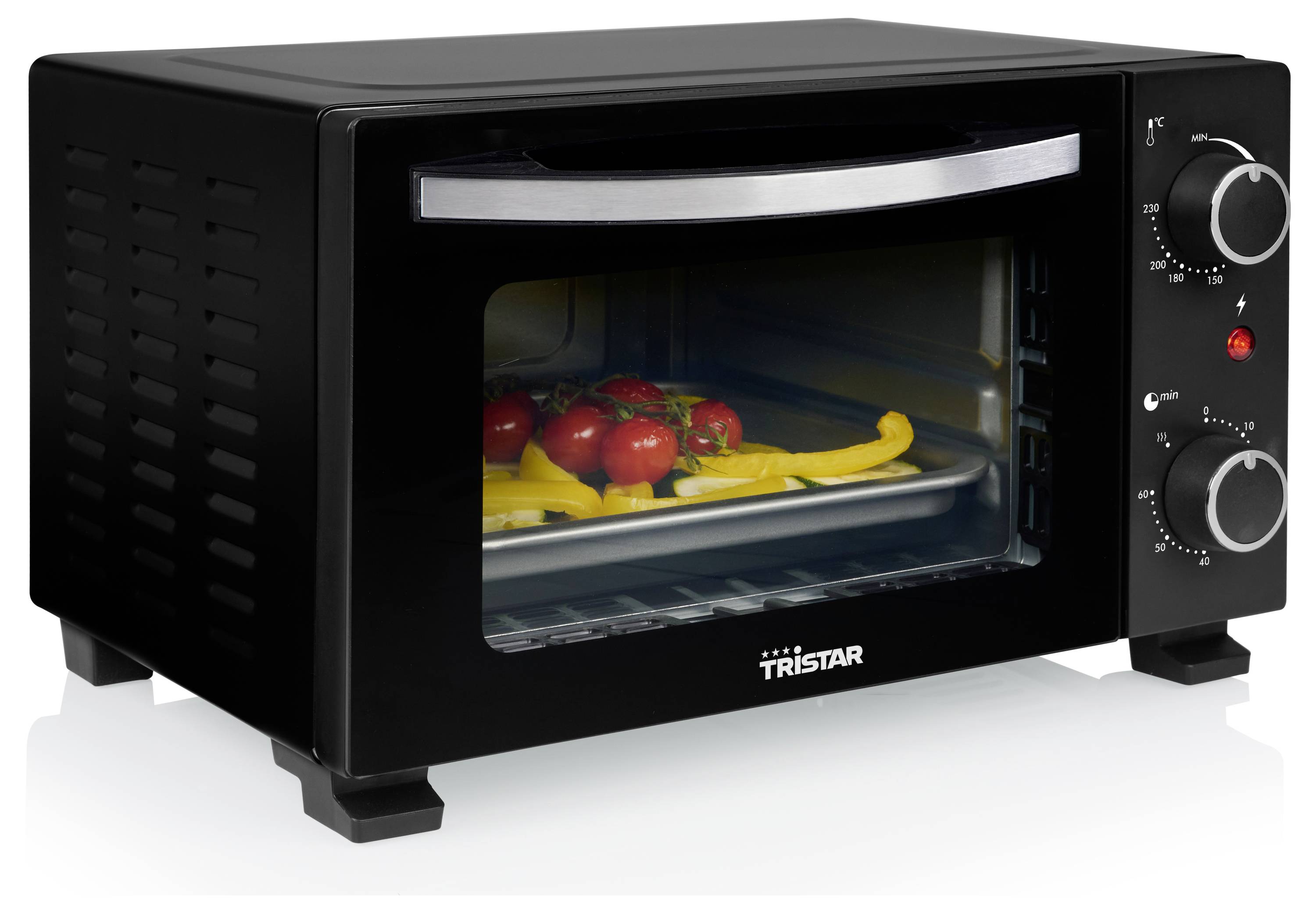 A small black oven with chrome handle and control knobs. Inside, a baking tray holds chopped red and yellow peppers and tomatoes.