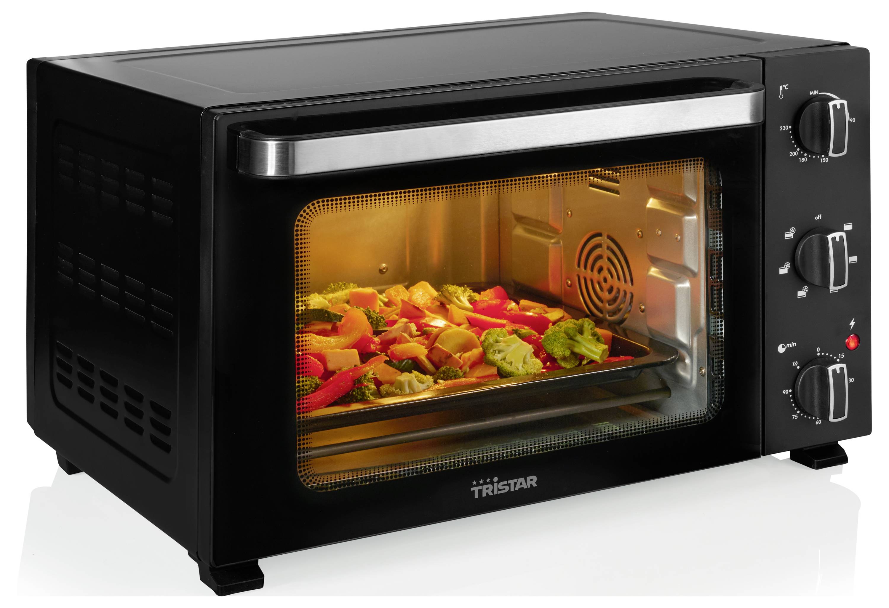 A black countertop oven with a silver handle is baking vegetables, visible through a glass door. Control knobs are on the right side.
