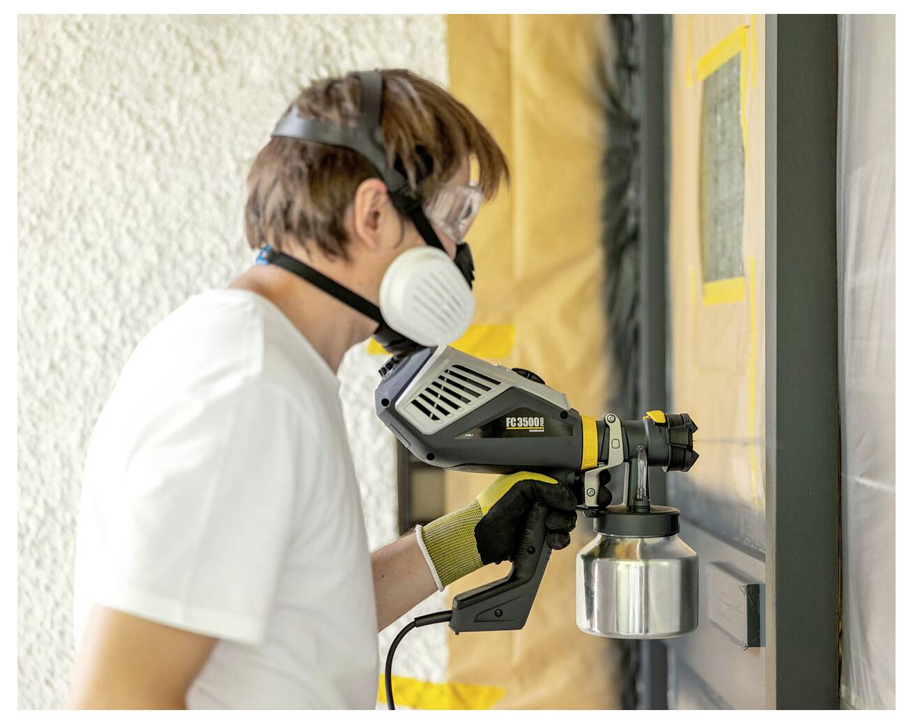 A person wearing a respirator mask and gloves uses a spray paint gun to apply paint to a masked-off door.