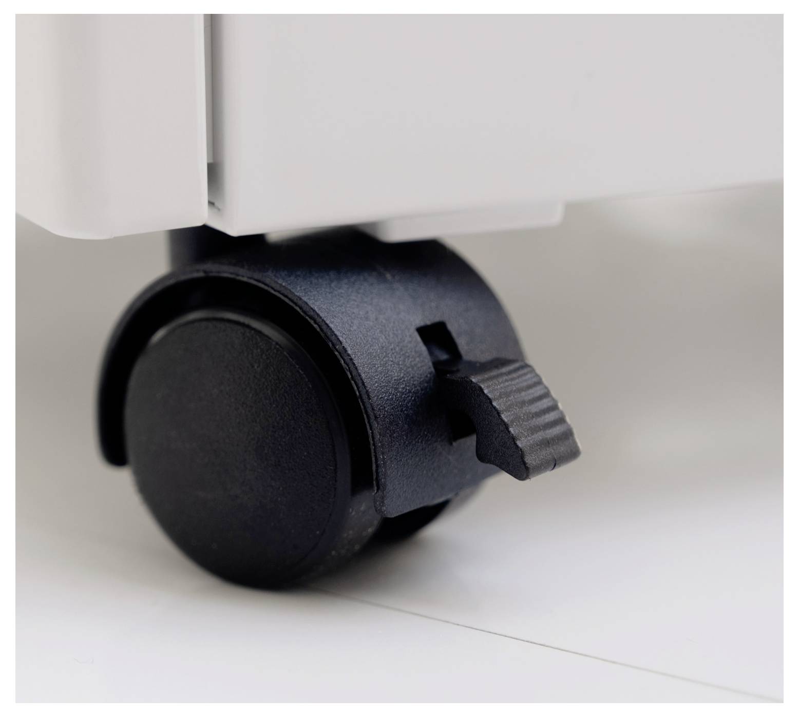 Close-up of a black caster wheel with a brake attached to a piece of furniture or equipment on a smooth white surface.