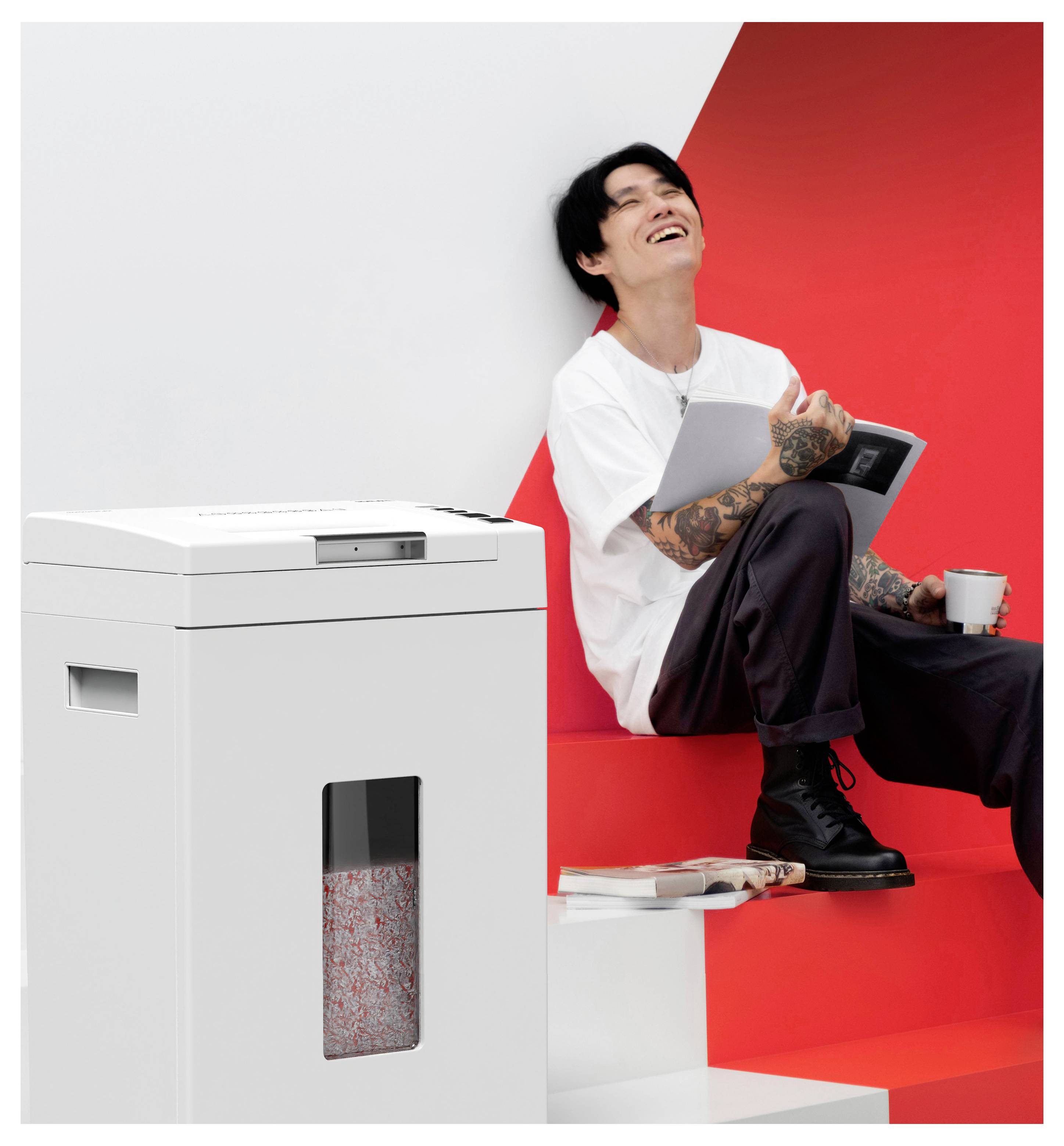 Person sitting on red steps, holding a tablet and cup, smiling. White paper shredder with visible shredded paper nearby.