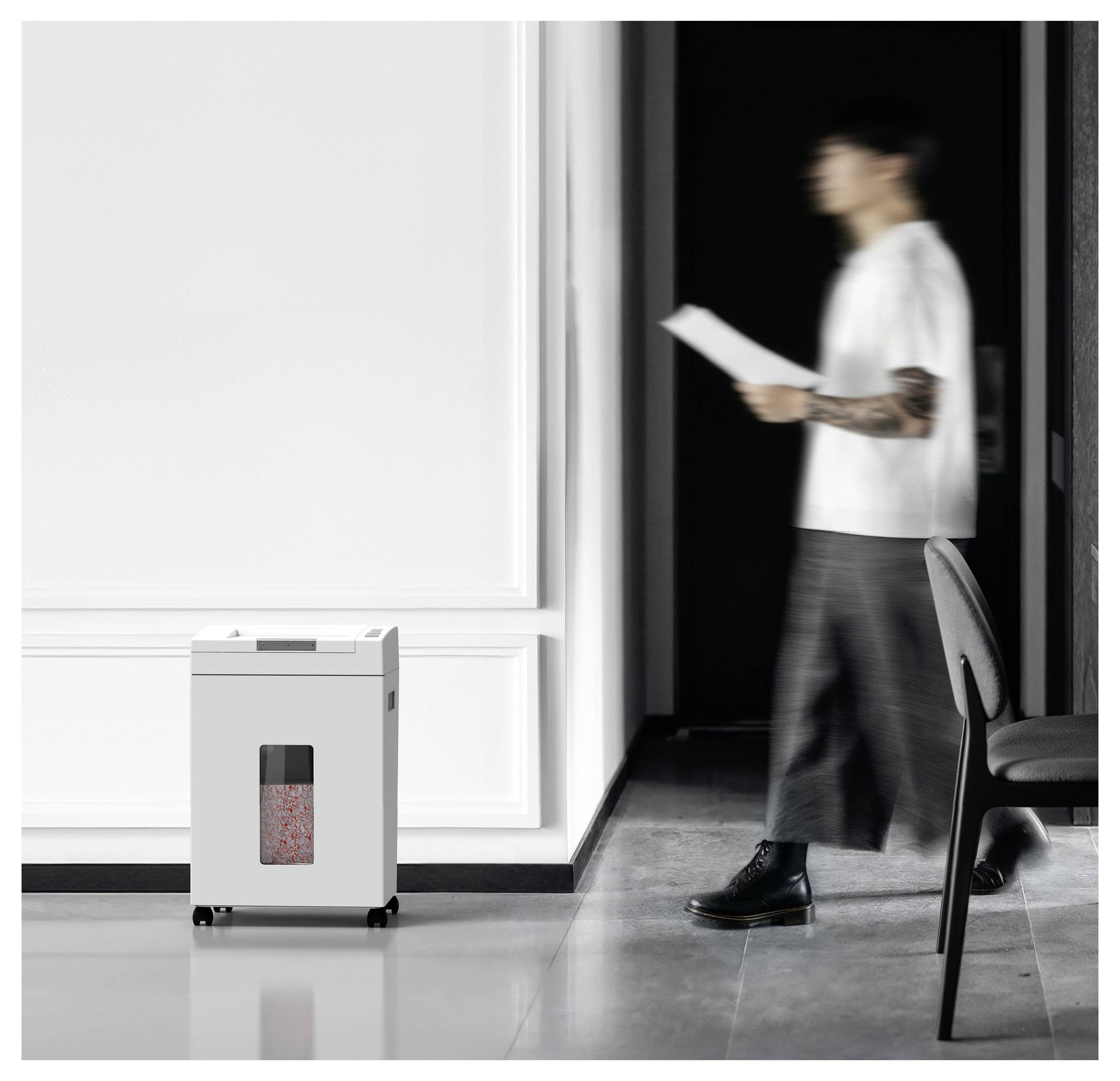 A person walks past a white paper shredder with shredded paper visible inside, placed in a modern office hallway.
