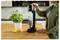 A person uses a soda maker on a kitchen counter next to a potted basil plant and two lemons. The machine is carbonating water in a bottle.