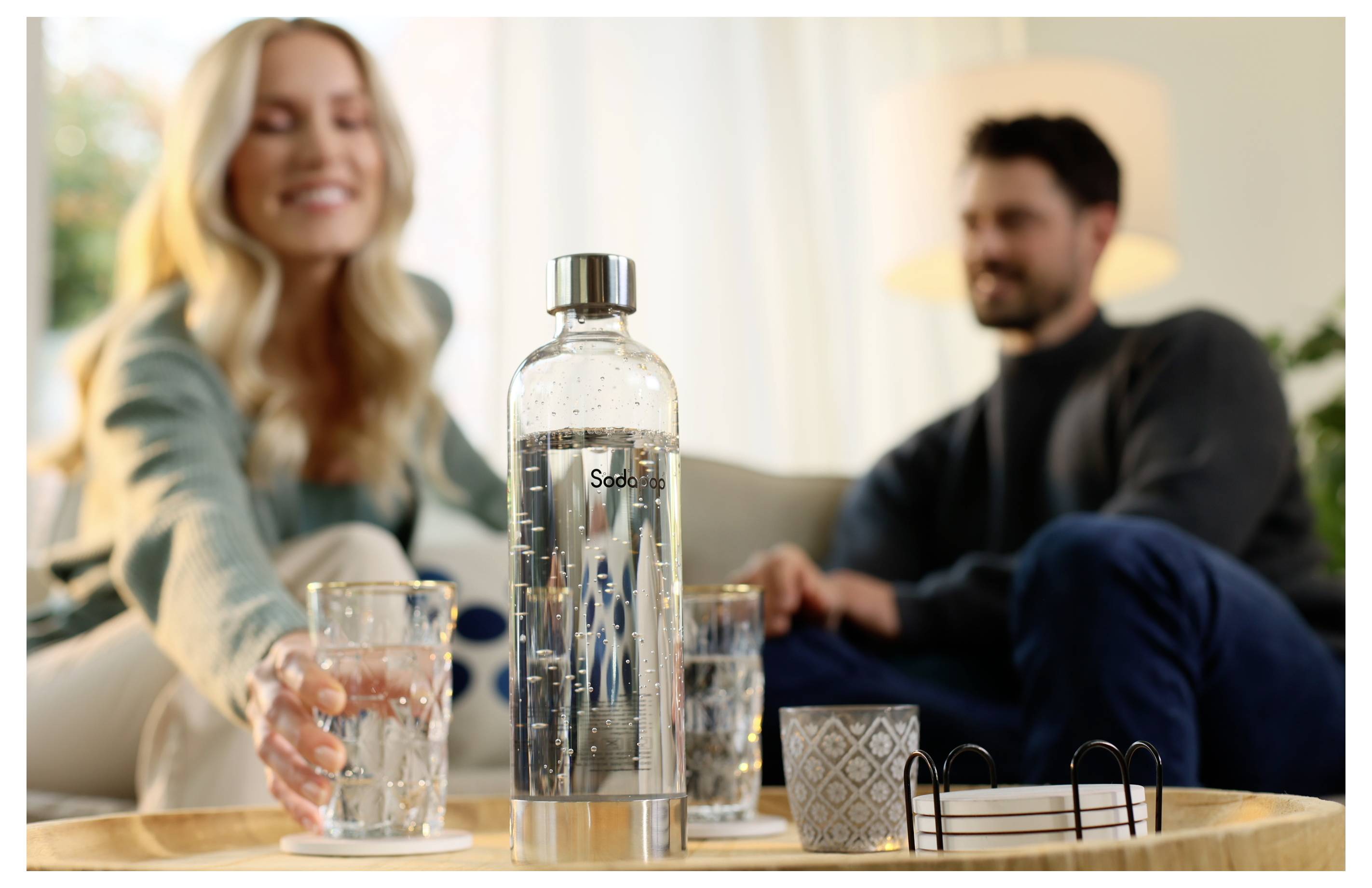 A woman handing a glass of sparkling water to a man on a tray with a SodaStream bottle and decorative items in a cozy living room.