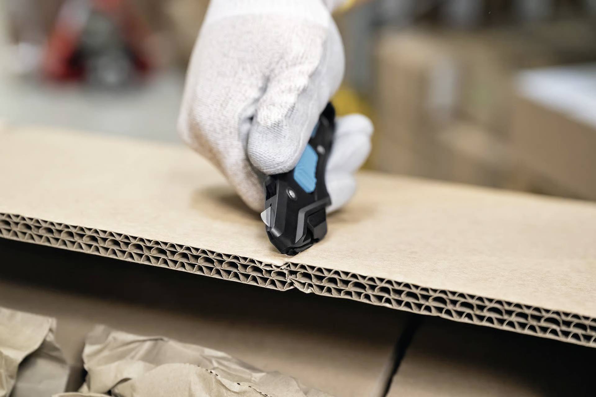 A gloved hand uses a utility knife to cut open a cardboard box, revealing brown packing paper inside, in a warehouse setting.