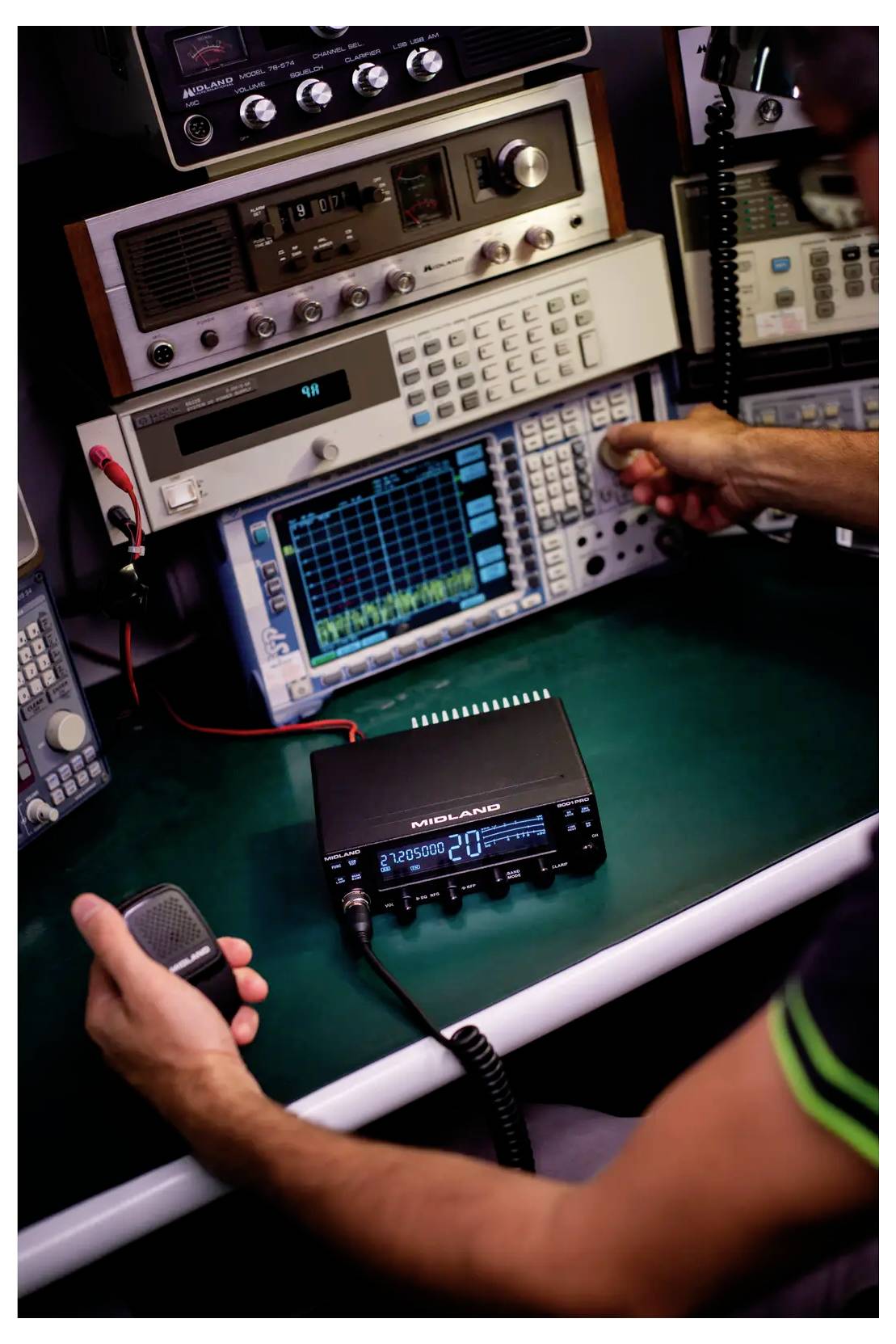 A person operates a radio communication setup, adjusting dials on equipment and holding a microphone. Various screens display waveforms.