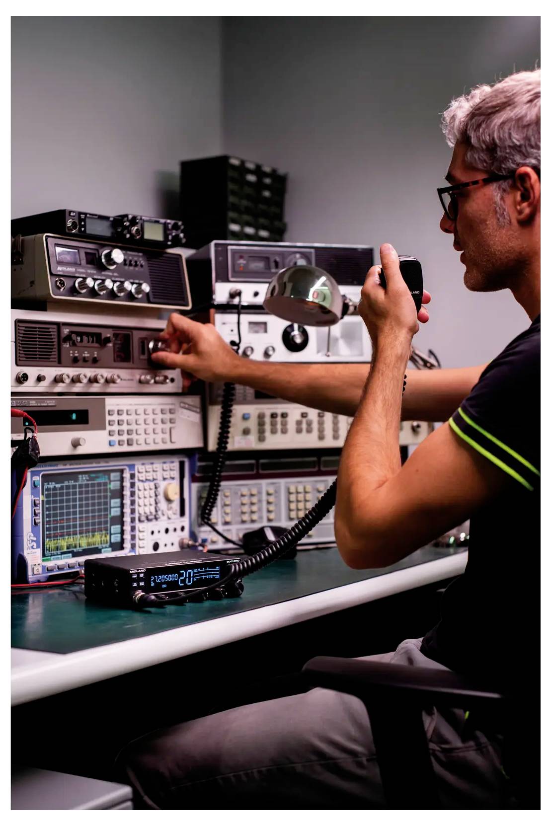 A person using a handheld radio microphone operates various radio communication equipment in a control room with monitors and dials.