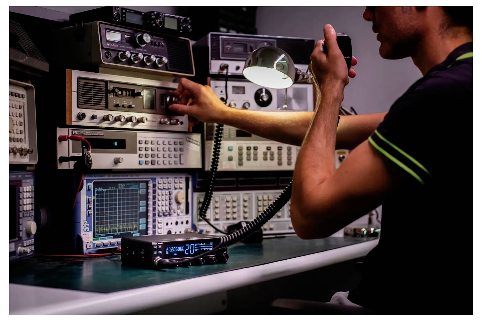 A person operates radio equipment in a communication station, adjusting knobs and speaking into a microphone, surrounded by electronic devices.