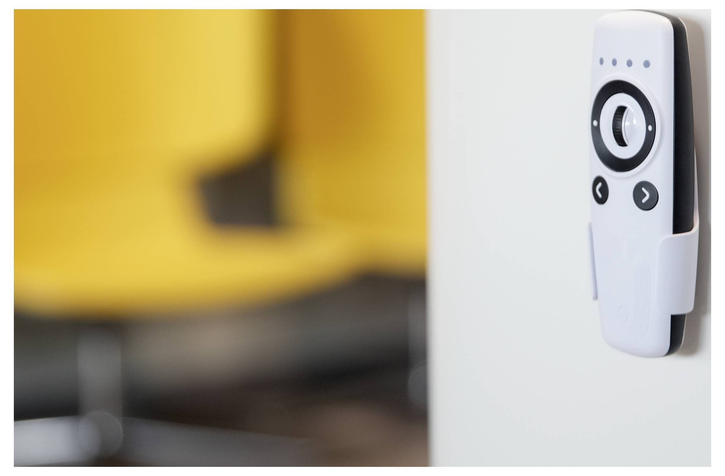 A wall-mounted remote control with a circular dial and buttons, partially blurred background showing yellow chairs.