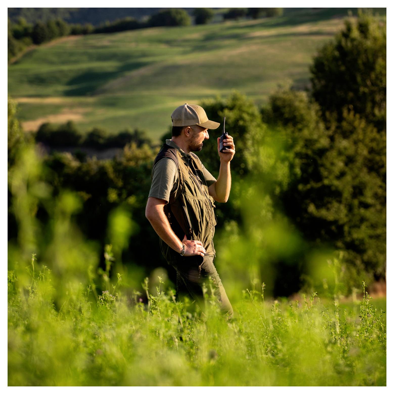 A person in outdoor clothing stands in a grassy field, holding a walkie-talkie, with rolling green hills and trees in the background.